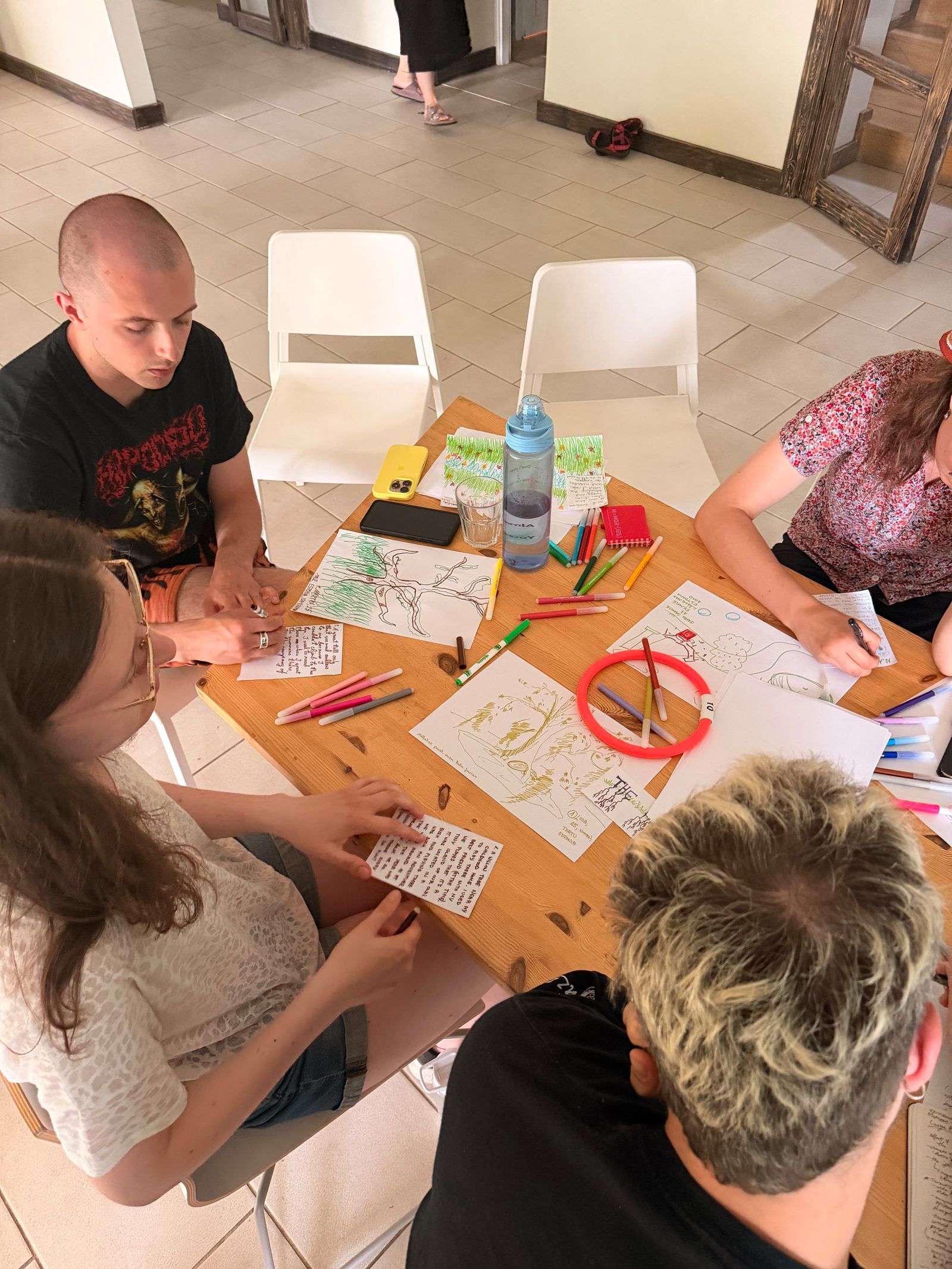 Overhead view of a small round table with four participants drawing, phones and coloured pens scattered between the sketches