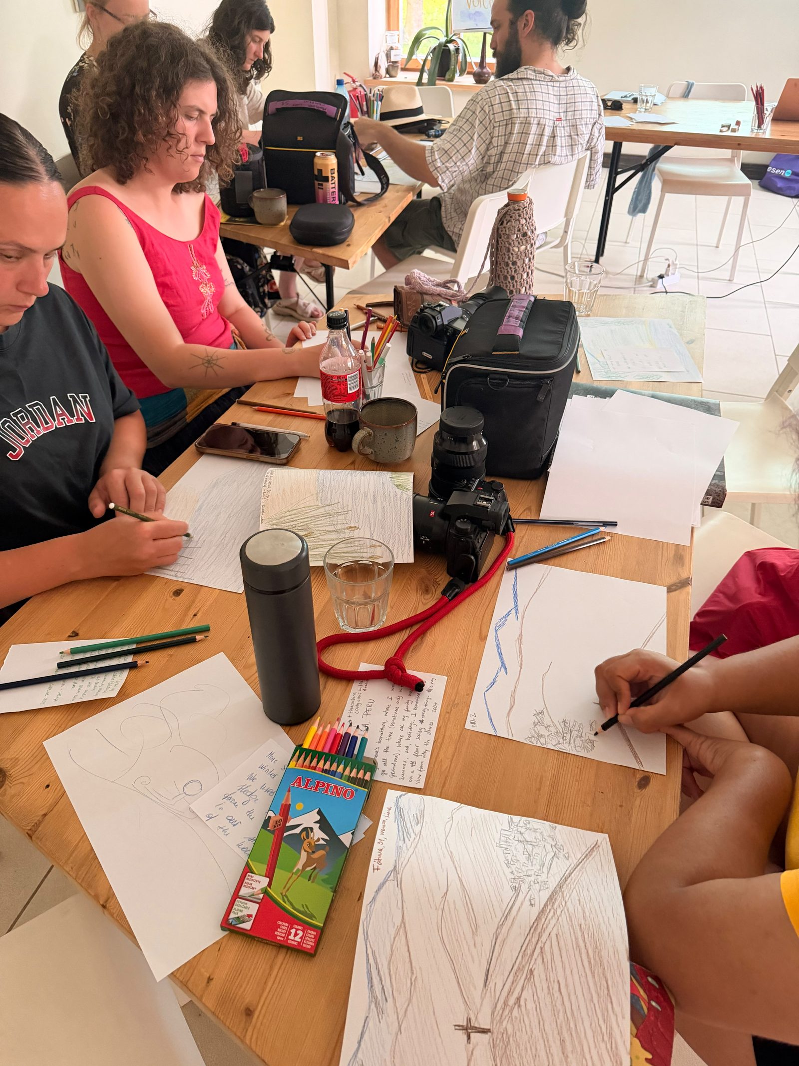 A long table crowded with participants drawing, pencils, and finished sketches of trees, rivers and mountains